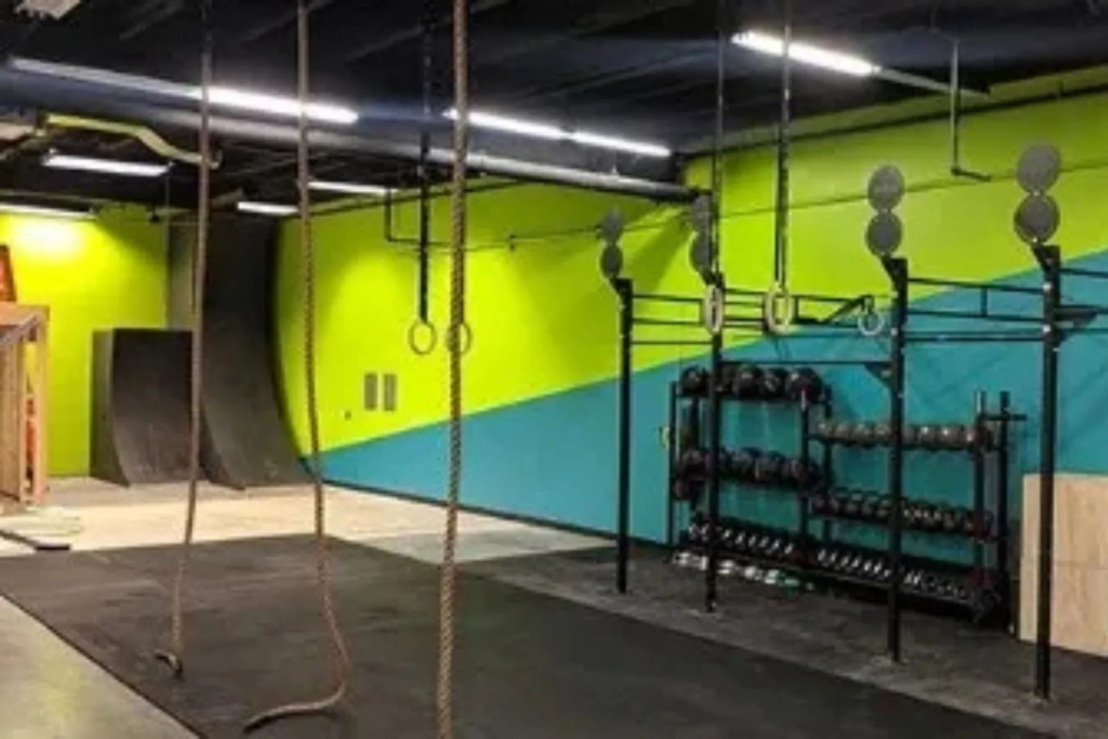 A functional fitness or CrossFit gym with black rubber gym flooring. The floor provides a durable and protective surface under a large rig, climbing ropes, a dumbbell rack, and other equipment, all set against a bold green and blue painted wall.
