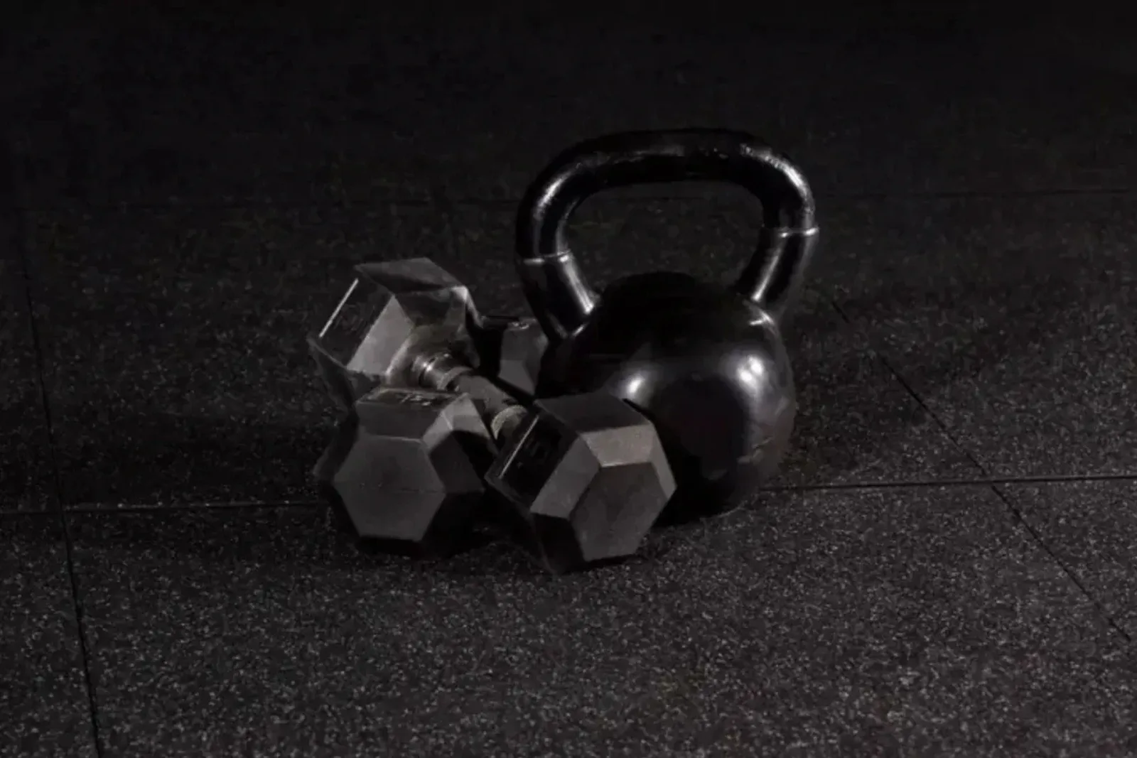 A close-up of a kettlebell and several dumbbells resting on black speckled rubber gym flooring. The durable rubber floor tiles protect subfloors from heavy weights and provide a non-slip surface for training.