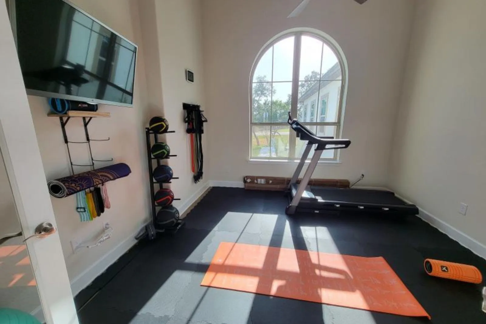 A bright home gym with natural light, featuring black interlocking rubber floor tiles. The durable rubber gym mats provide a protective surface for a treadmill, workout accessories like medicine balls and resistance bands, and a yoga mat placed on top.