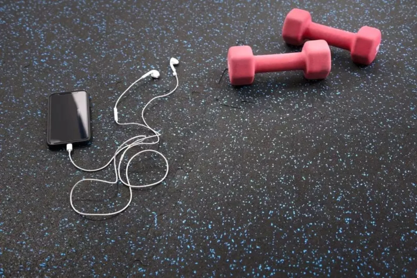 A vibrant commercial gym or fitness studio with rubber gym flooring in a gray and light-colored pattern, accented with a blue stripe. The space features a heavy punching bag, kettlebells, medicine balls, and brightly colored workout equipment against a ge