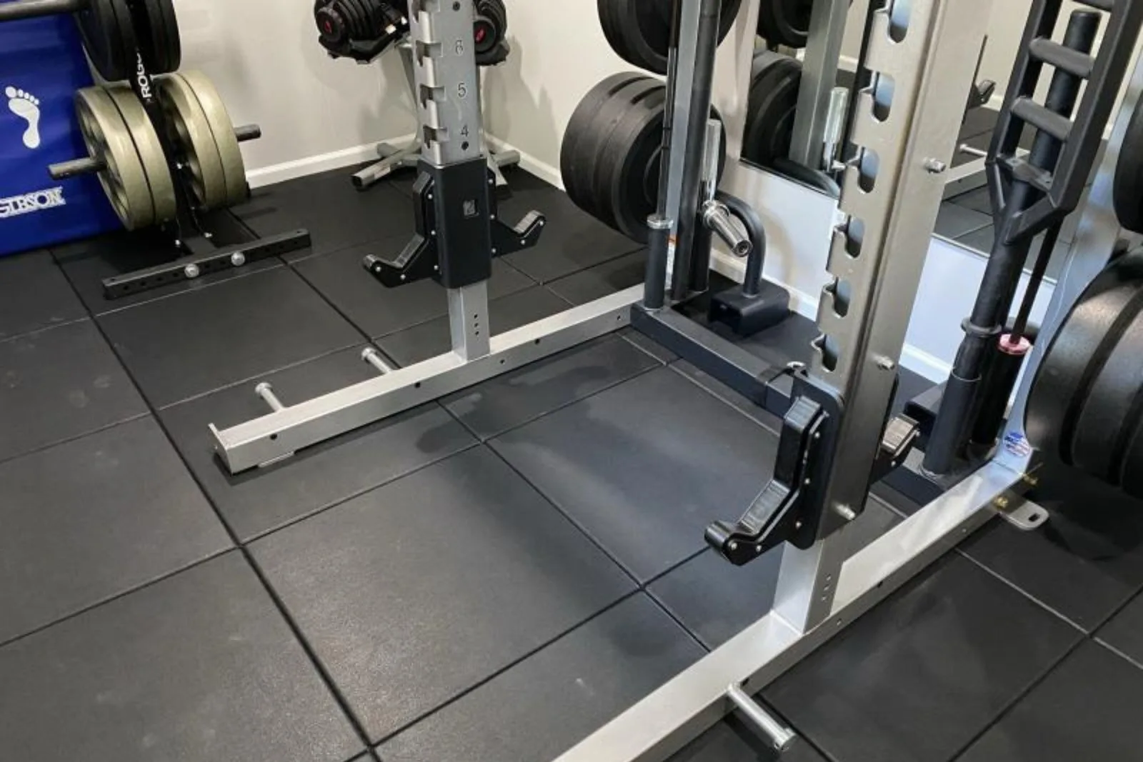 A close-up of a home gym's floor, showcasing black interlocking rubber tiles. The durable gym flooring provides a protective and stable base for a power rack and organized weight plates, ensuring the subfloor is safe from drops and heavy equipment.