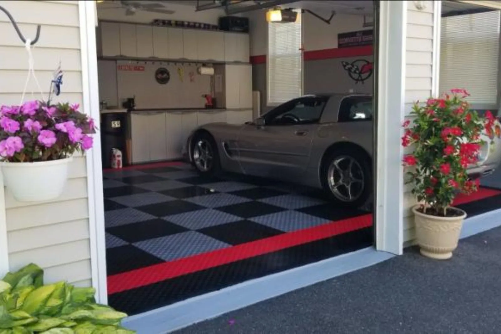 A custom Corvette garage featuring black and grey interlocking floor tiles, a red border, and extensive white garage cabinets.
