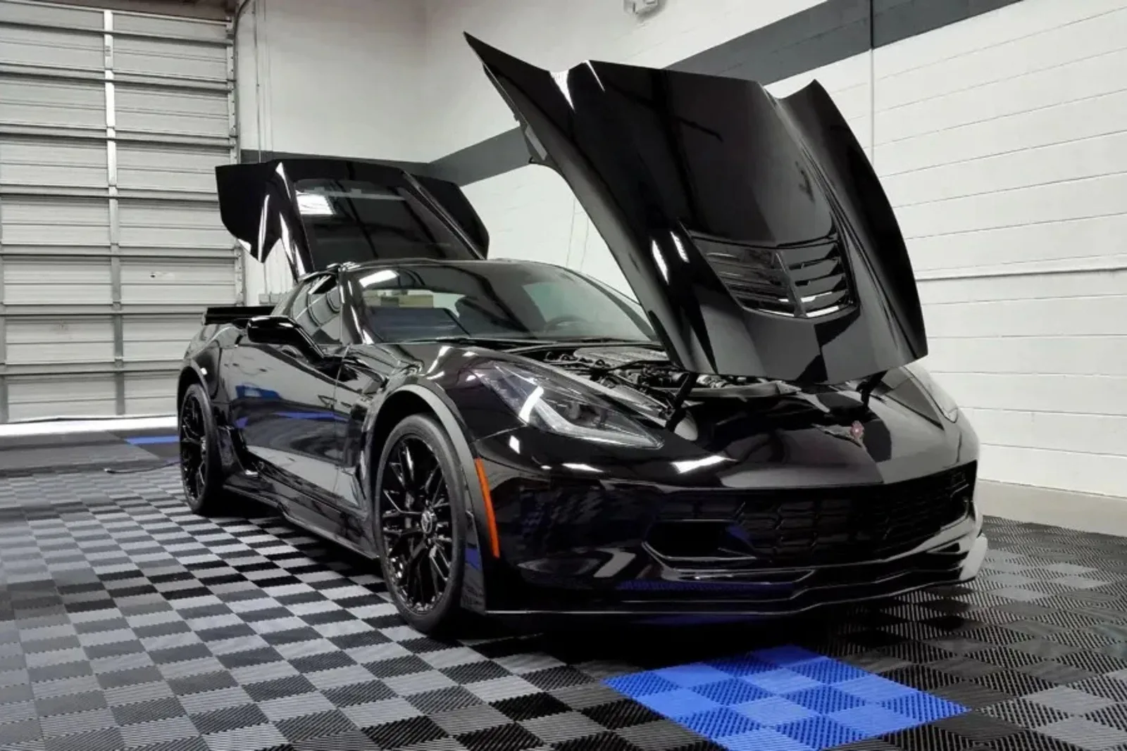 A black Corvette on interlocking garage tiles in a black, grey, and blue checkerboard pattern, showcasing a custom garage showroom.