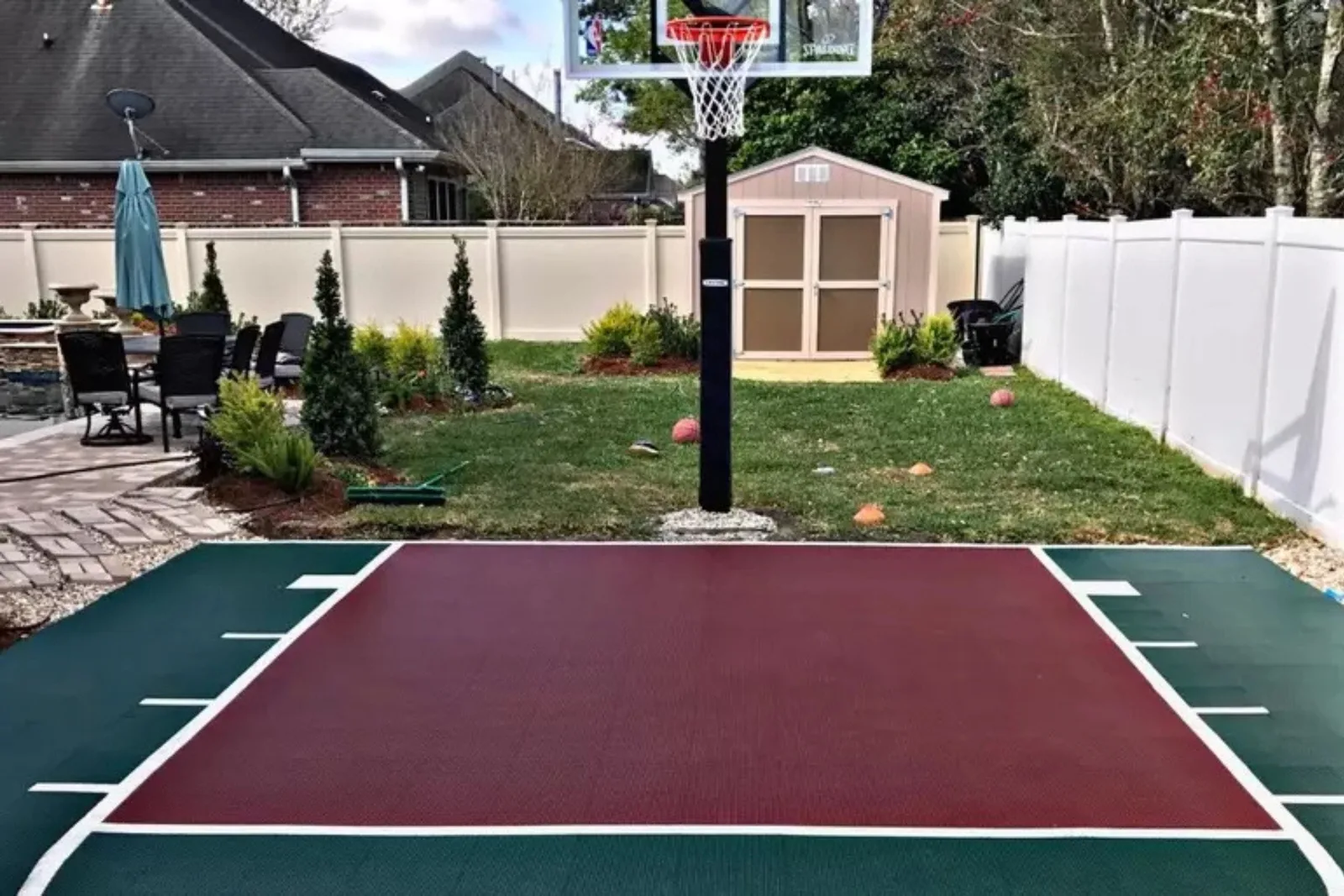 Outdoor basketball court kit with red and green interlocking court tiles, creating a multi-sport playing surface in a residential backyard with a shed and patio.