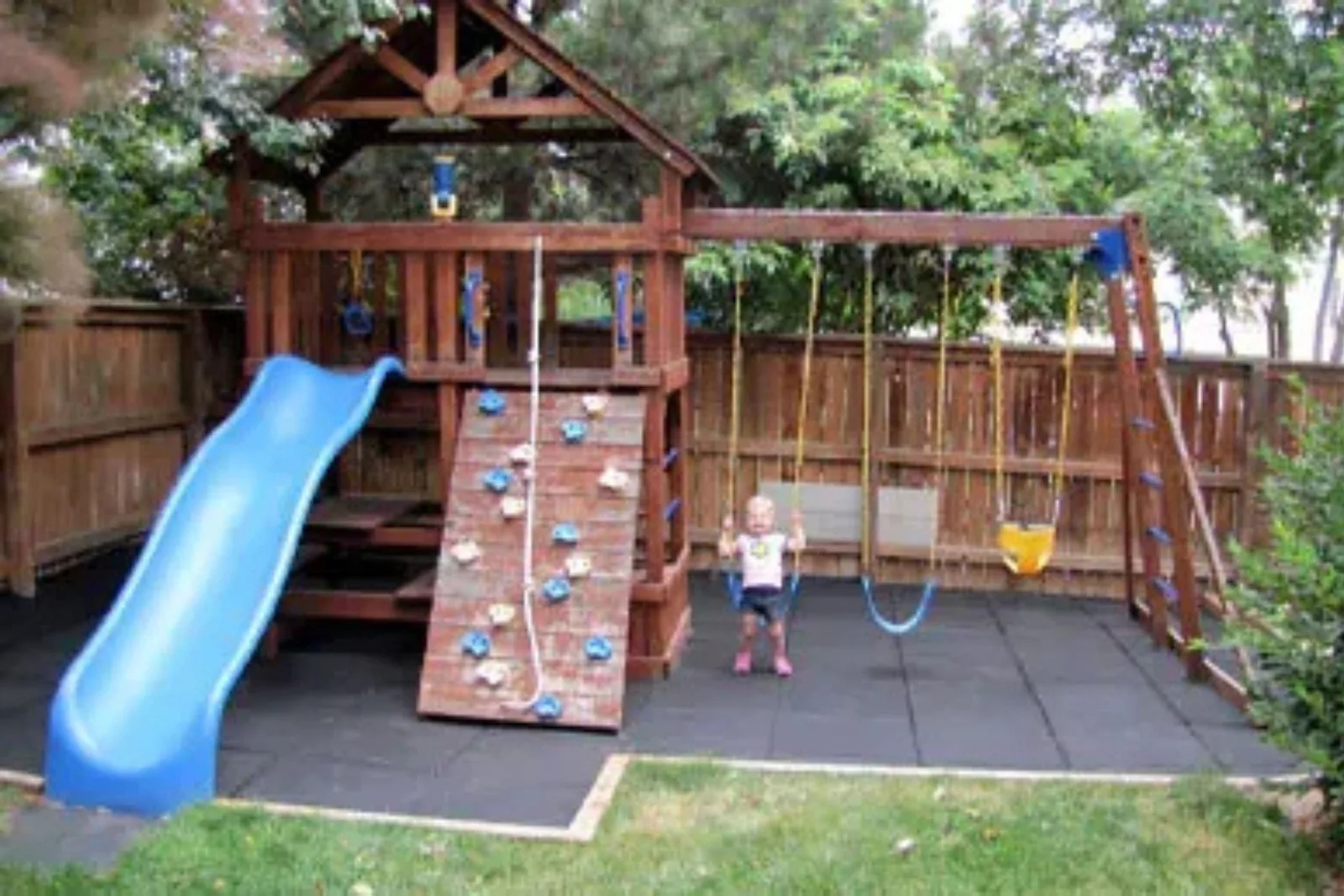 Durable rubber playground tiles in a backyard, creating a safe and cushioned surface under a wooden playhouse with a slide and swings.
