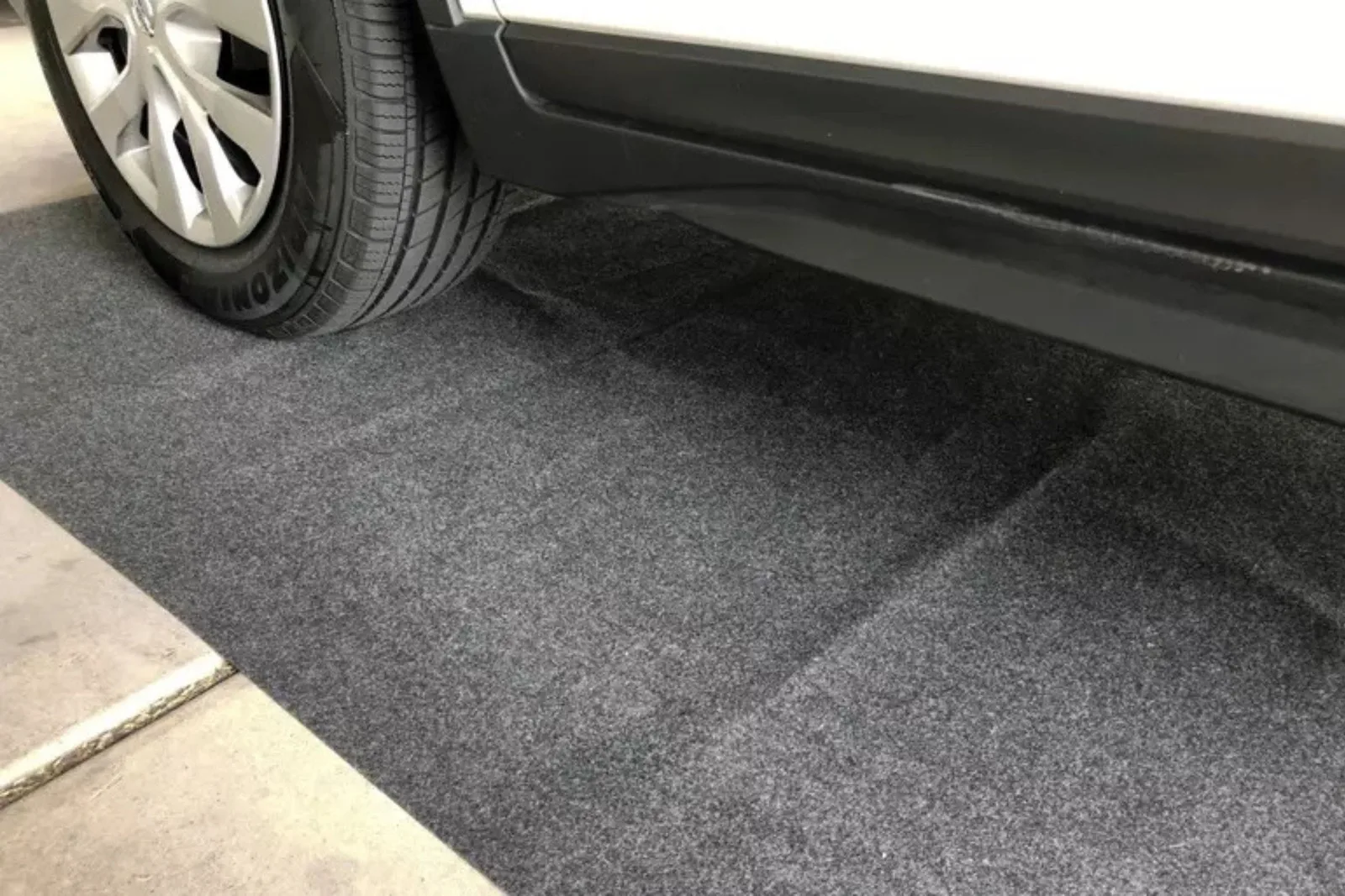 The wheel of a white car is parked on a gray carpet garage mat. This durable garage runner is designed to absorb liquids and protect the concrete floor from spills, drips, and corrosive fluids.
