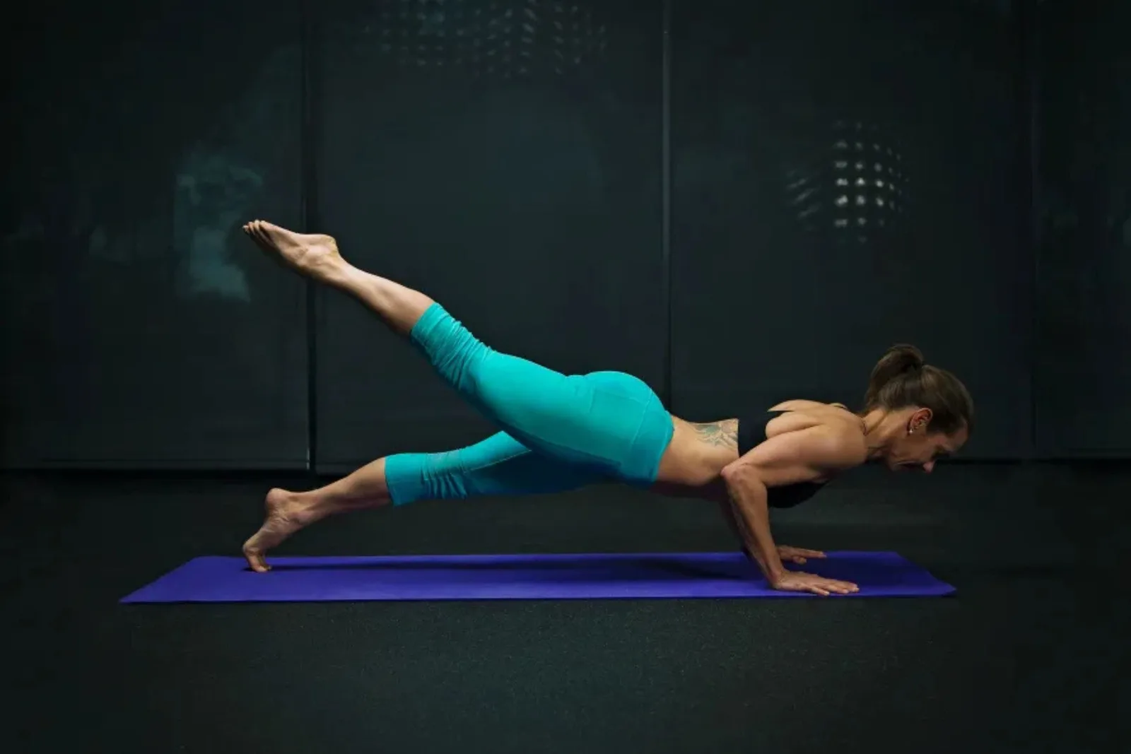 Durable black rubber gym flooring Woman performing a single-leg push-up variation on a purple yoga mat over durable black rubber gym flooring, a protective surface for fitness.