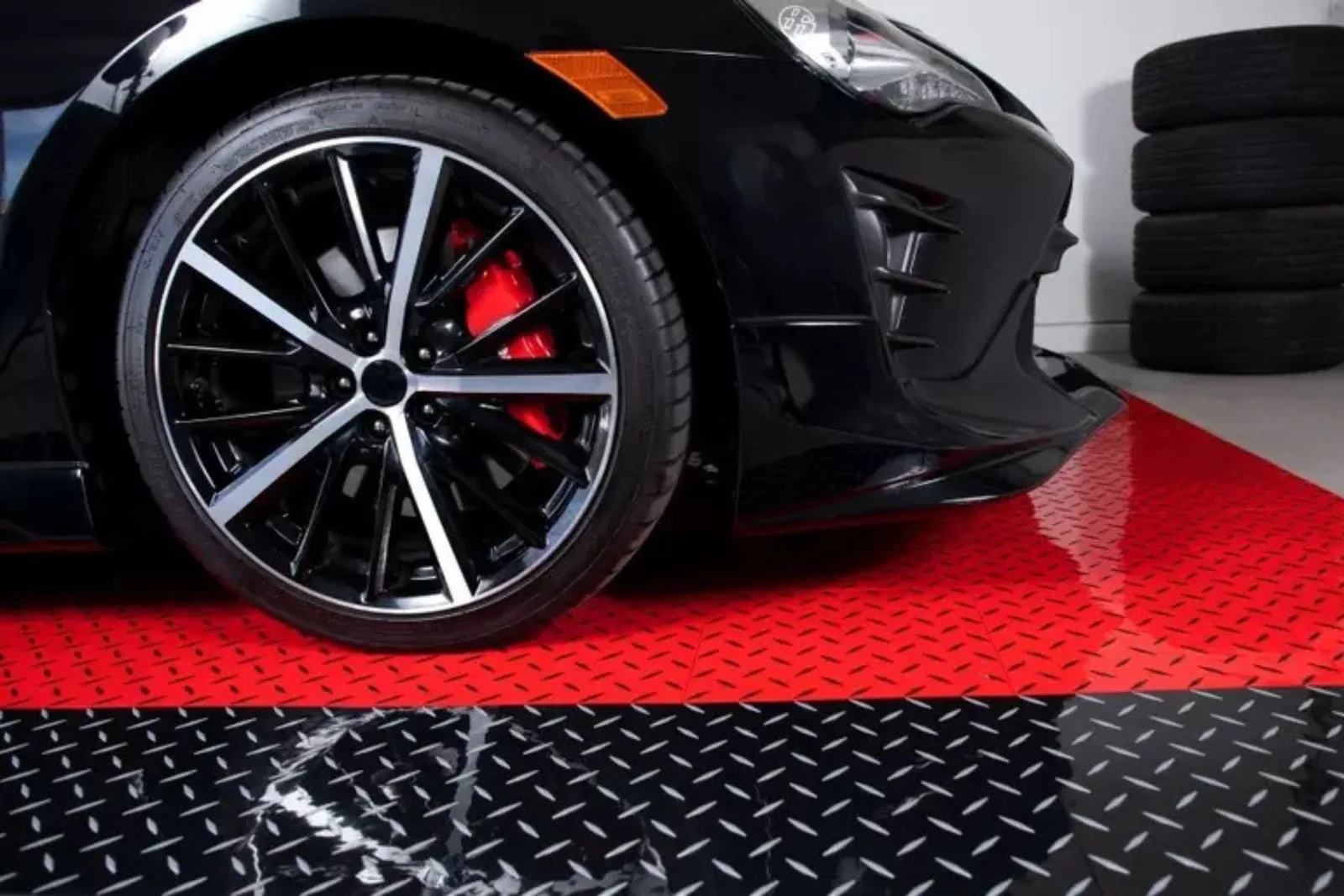 Durable diamond-plate garage tiles Close-up of a black sports car on durable red and black diamond-plate plastic garage tiles, showcasing a high-performance and stylish flooring solution.