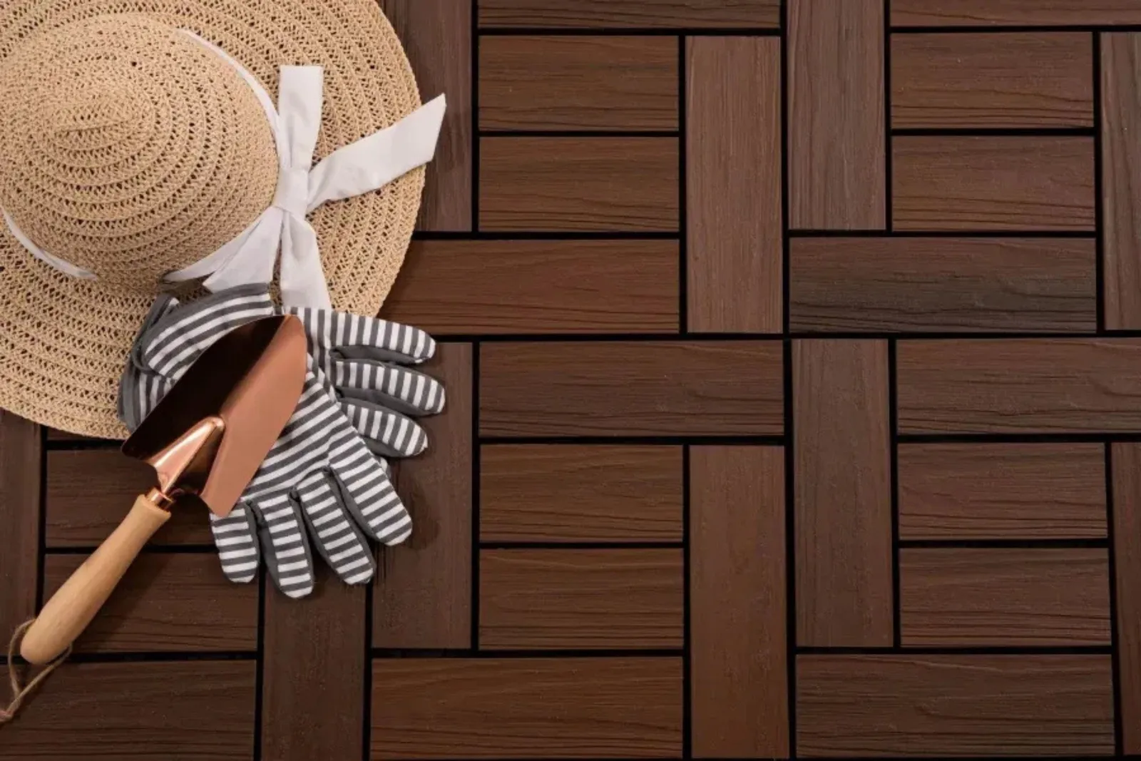 Composite deck tiles in a dark brown 6-slat wood grain pattern, creating a stylish and low-maintenance outdoor flooring solution for a patio or garden, shown with a sun hat and gardening tools.