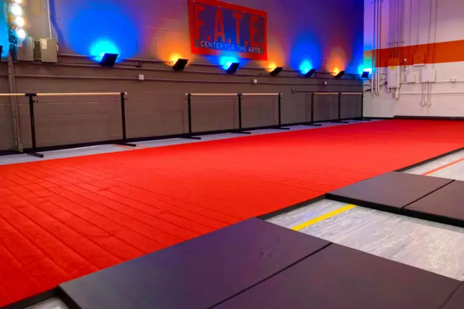 Carpet-topped foam gymnastics mat in bright red creates a long tumbling strip in an arts center, flanked by mirrored walls, dance barres, and black landing mats.